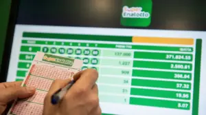Mani che compilano una schedina del SuperEnalotto davanti a uno schermo con i risultati e le quote della vincita