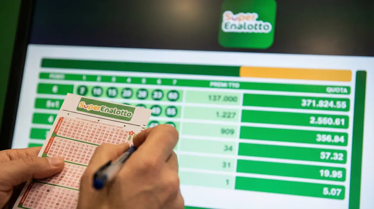 Mani che compilano una schedina del SuperEnalotto davanti a uno schermo con i risultati e le quote della vincita